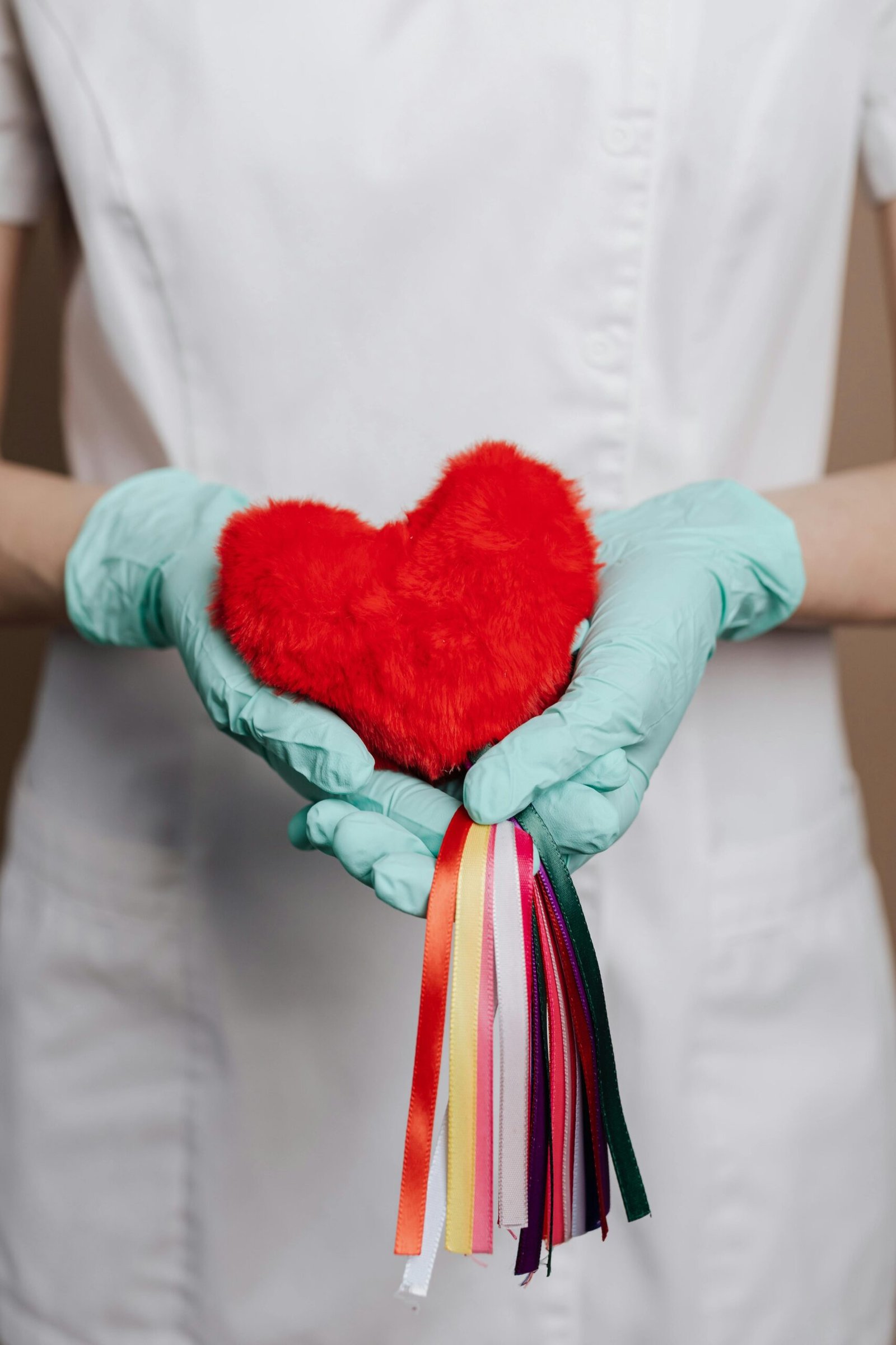 A healthcare worker with gloves holds a red heart-shaped object adorned with colorful ribbons, symbolizing care and love.
